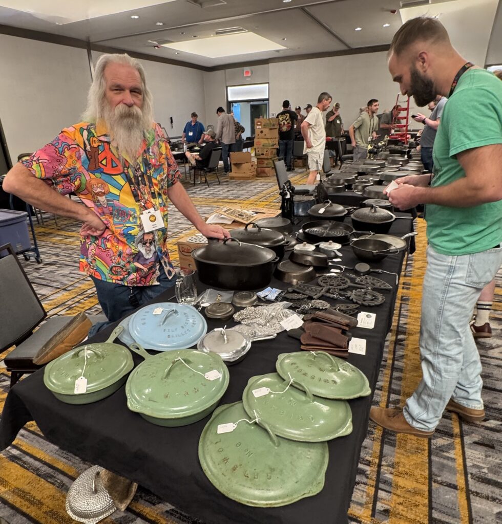 Man at table with many pieces of cast iron cookware at the swap meet of the at the cast iron convention. 
