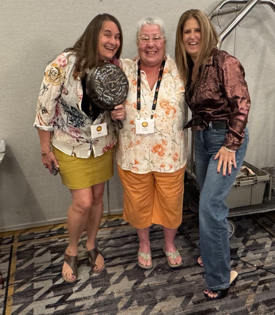 3 ladies with one holding a marked-up cast iron pan at the cast iron convention. 